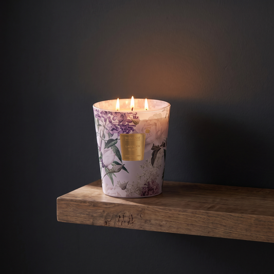 A three-wick candle in a clear glass jar with a light pink floral print and a gold label, sitting on a rustic wooden shelf against a dark gray wall.