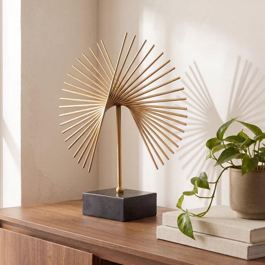 A decorative table sculpture with a gold-toned fan-shaped metal sculpture on a black rectangular base, styled on a wooden console table next to a potted green plant and stacked books.