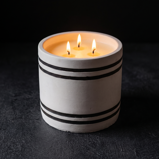 A three-wick soy wax candle in a cylindrical off-white ceramic holder with black horizontal stripes, shown lit on a dark textured surface against a dark background.