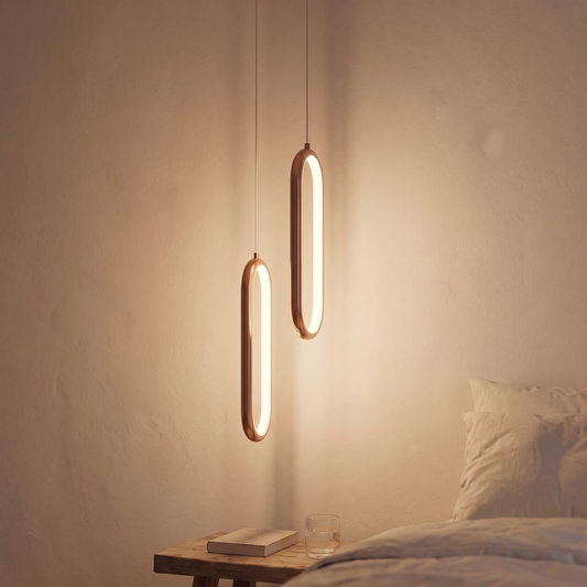 Two modern vertical oval pendant lights with warm white glowing LED strips and light wood frames, hanging in a bedroom above a bedside table next to a bed.