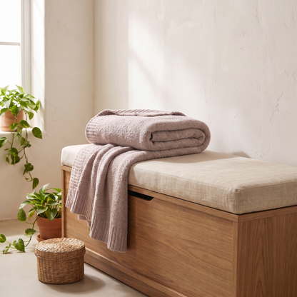 A folded dusty pink knit blanket and a draped matching throw are displayed on a wooden storage bench with a beige upholstered seat in a bright, sunlit room with potted plants.