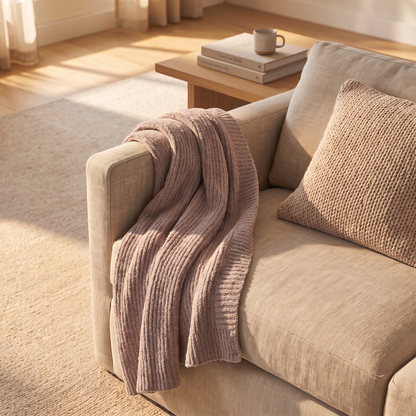 A cozy beige sofa in a sunlit room with a ribbed dusty pink throw blanket draped over the arm and a matching knit pillow, with a small wooden side table and books in the background.