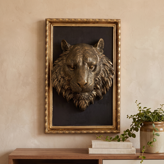 A framed wall art piece featuring a sculpted lion head in metallic bronze tones against a dark background, hung on a beige wall above a wooden console table with books and a potted plant.